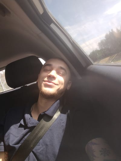 A selfie is taken from inside a car, looking toward the sunroof. The driver, a young man with a light beard and short hair, is smiling with his eyes closed. He is wearing a dark blue polo shirt and a silver chain. A seatbelt is diagonally across his chest. The interior of the car is dark gray. Outside the car, through the sunroof and windows, a bright sky with some clouds is visible, along with trees and a road. The photo appears to be taken during daylight. The provided crops do not offer additional context about the city of Cherkasovo, Russia.