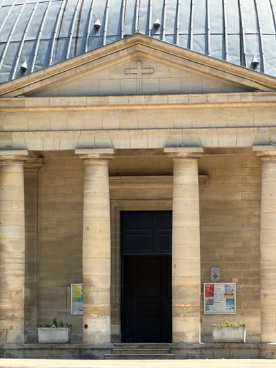 The image shows the facade of a building, likely a church, in Courbevoie, France. The building features a large, rounded, metal-clad roof with visible seams and several conical elements protruding from it. Below the roof, a triangular pediment with a sculpted cross is visible, resting on a stone entablature. The entablature is supported by a series of large, fluted columns made of pale stone. Between the columns, there are sections of a stone-block wall. A large, dark-colored double door is centered in the facade, accessible via a short set of steps. To the left of the entrance, a white planter box contains a few small green plants. A framed notice board with multiple posters is mounted on the wall to the left of the door. To the right of the door, another, smaller notice board with red lettering and a symbol is visible. A second planter box with sparse greenery is located to the right of the door, closer to the edge of the frame. The overall lighting suggests it is daytime, with shadows cast by the architectural elements.