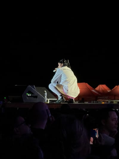 A woman is squatting on a stage, holding a microphone and wearing a white jacket with "HUMAN" written on the back. She has black hair in a ponytail and is wearing white tights and black boots. In the background, there are orange tents with the word "AMBAR" visible on one of them. The stage is set up in an outdoor venue, possibly a music festival, with a crowd of people in the foreground. The event appears to be taking place in Rivas-Vaciamadrid, Spain.
