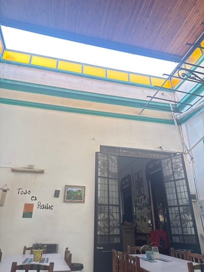 This image captures a brightly lit, open-air or semi-covered patio space, likely belonging to a hostel or cafe in San Miguel de Tucumán, Argentina. The scene suggests daytime with ample natural light.
On the left, a slatted wooden ceiling or awning runs along one side, contrasting with a large, bright window opening featuring rectangular yellow stained-glass panels framed by a dark, possibly wrought-iron, railing with decorative scrolls. The ceiling in other parts of the patio is white, with some exposed piping and a security camera mounted high on the wall.
The main wall of the patio is cream-colored with vertical light green/teal stripes. A prominent feature is a large, dark double door with ornate grilles, likely leading into an indoor area. Through the glass panels of this door, several signs are visible, including "A LA BURDO HOSTEL", indicating the establishment's nature. Other text seen through the glass includes "IF NOT NOW WHEN?", "BOTIQUIN", and "PRIMEROS AUXILIOS" (First Aid Kit, First Aid).
The cream wall is decorated with various items: a framed landscape painting, a smaller dark square art piece, and the hand-painted Spanish phrase "Todo es Posible" (Everything is Possible) in a flowing script. A green flyer or poster is affixed near this text, and a macrame wall hanging is visible further down. A long, cylindrical light fixture or decorative element is also mounted on the wall.
To the right and in the foreground, several wooden tables and slatted chairs are arranged, suggesting a dining or lounging area. One table holds a small potted plant and a tissue box, adding a touch of domesticity. The space appears inviting and functional, offering a blend of rustic charm and artistic elements.