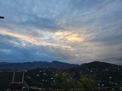 A panoramic view shows rolling hills dotted with small houses under a cloudy sky. The sun filters through a gap in the clouds, casting a warm glow. The terrain is uneven, with some areas appearing densely wooded. The sky is mostly gray and blue, suggesting either a sunrise or sunset.