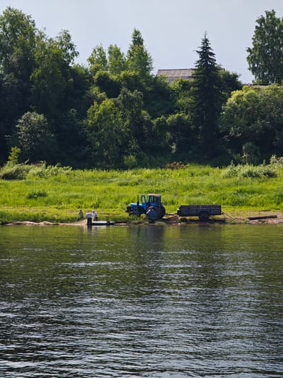 In Усть-Кемь, Russia, on a sunny day, a blue tractor is parked on a grassy bank next to a river. Attached to the tractor is a dark trailer. Several people are near the river's edge, some in a small inflatable boat, and others standing on the bank. One person is using a fishing net. The river is calm with ripples reflecting the green trees and blue sky. Behind the bank, a dense forest covers the landscape, with a few houses visible through the trees on a higher elevation.