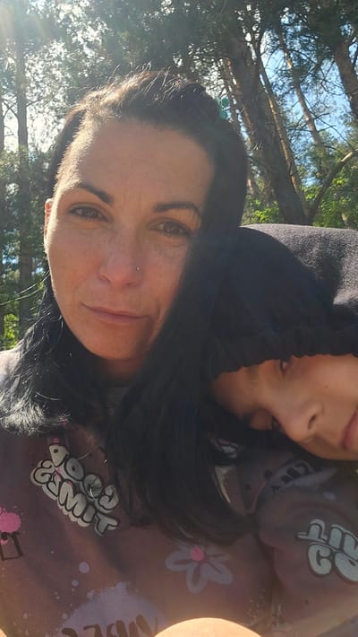 In Dolni Pasarel, Bulgaria, a selfie captures two individuals outdoors, nestled amongst tall trees under a bright sky. The person in the foreground, appearing to be an adult woman with dark hair, faces the camera with a gentle smile and a nose piercing. Sunlight dapples through the leaves, illuminating her face and casting shadows. Her head is turned slightly, and her dark hair cascades over her shoulder. Resting against her is another person, a child with dark hair, their face partially obscured by the hood of a dark garment and their head laid on the adult's shoulder. They appear to be relaxed, with their eyes closed or looking down. The clothing of both individuals features graphic designs, with the adult's garment displaying stylized text that reads "Good Times" and floral motifs. The background is dominated by the natural scenery of trees, with glimpses of blue sky peeking through the canopy. The overall impression is one of warmth, affection, and shared outdoor time.