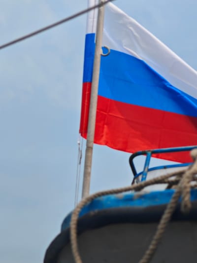 The image shows a Russian flag waving against a pale blue sky. The flag is composed of three horizontal bands of white, blue, and red. A flagpole, made of metal, is visible on the left side of the flag. In the foreground, the blue railing and a thick rope of a vessel are out of focus, suggesting the photograph was taken from a boat or ship. There is no visible indication of the city of Peschanka, Russia.