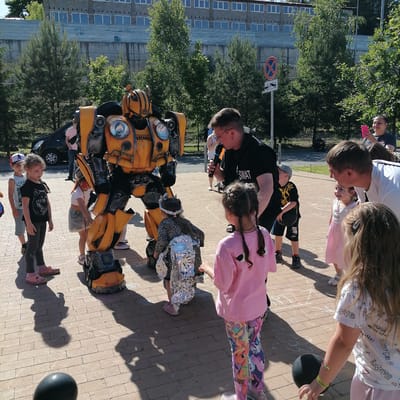 An outdoor event is depicted on a sunny day in Трубачеевка, Russia, featuring a person in a Bumblebee Transformer costume, an entertainer, and a crowd of children and adults. In the center-left, a large, yellow and grey Bumblebee Transformer costume stands on a paved brick surface, facing generally right. To its immediate right, an adult male entertainer, with short hair and glasses, wearing a black t-shirt with "SWAT" printed on it, holds a black microphone to his mouth, directing his attention towards the children. Numerous children, ranging from toddlers to early elementary age, are gathered around the Bumblebee costume and the entertainer. To the left of Bumblebee, a girl in a black t-shirt with a wolf graphic and dark leggings stands. Behind her, a boy in a white tank top and a red and white baseball cap observes. Close to Bumblebee's right side, a girl with red face paint on her cheek, wearing a white crop top and denim shorts, looks towards the costume. A girl in a pink t-shirt and colorful My Little Pony leggings stands with her back to the camera on the right side of the frame. Another child, wearing a leopard print dress and a silver foil backpack, is positioned near Bumblebee's feet. A small boy in a black t-shirt and shorts holds a black ball while looking up at the entertainer. To the far right, an adult male in a white t-shirt leans towards a smiling small girl in a pink Hello Kitty dress, who has light face paint. In the foreground right, a child with long hair holds a black balloon. Two black balloons rest on the ground in the foreground. Faint white chalk drawings, possibly a game layout, are visible on the light brown/beige brick pavement. In the background left, a black car is partially visible. In the mid-ground right, a road sign assembly consists of a circular "No Stopping" sign (red circle, blue background, red diagonal line) above a rectangular sign depicting a tow truck and car, indicating a no-towing zone. A large, multi-story building with numerous square windows and a concrete facade occupies the far background. Several green trees, both coniferous and deciduous, are scattered throughout the background. The strong sunlight casts distinct, elongated shadows of the figures onto the paved surface, indicating a clear, bright day. In the background right, an adult female with dark hair tied back, wearing a