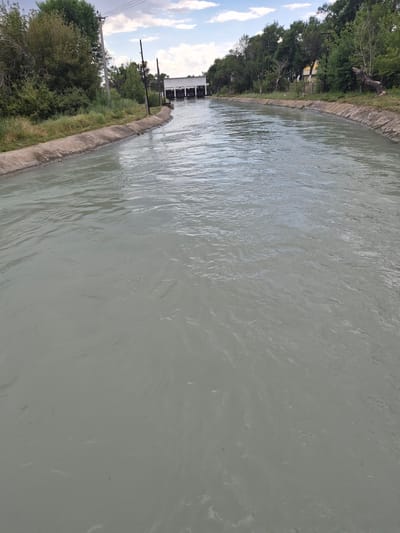 The image depicts a wide, straight canal filled with murky, gray water. The canal has concrete-lined banks covered with grass and vegetation on the left side. On the right side, the bank is also concrete-lined, with a rocky incline leading up to trees and some buildings in the distance. A large, white, utilitarian building with dark openings is visible at the far end of the canal, suggesting it might be a pumping station or sluice gate. The sky is partly cloudy with blue patches. Power lines are visible running along the left bank. The scene appears to be an engineered waterway within a rural or semi-rural area, possibly on the outskirts of the city of Tekeli, Kazakhstan, given the visual characteristics of the vegetation and architecture. There are no people or animals visible in the image.