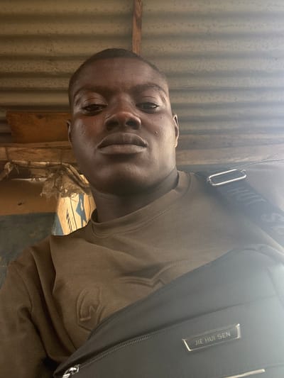 A young Black man is depicted in a medium-close-up selfie, looking directly at the camera with a neutral expression. He is wearing a long-sleeved, brownish-green or olive-colored shirt, which has an embossed "J" or similar symbol visible on his right sleeve. A black backpack or bag is slung over his right shoulder. The bag features a silver rectangular tag with the text "JIE HUI SEN" written vertically. Part of the bag's strap, equipped with a silver buckle, also displays faint, partially obscured text, possibly "SHUI".

The background suggests an informal, possibly semi-outdoor setting in Akodo, Nigeria. To the man's left, corrugated metal sheets, likely forming a wall or fence, are visible. Wooden planks and beams are also present, indicating a simple or rustic structure. Below his chin and towards the right, a light blue object with faint white lettering, possibly "WM" or "WIM," can be seen. The scene is brightly lit, suggesting it was taken during the daytime, with some glare evident.