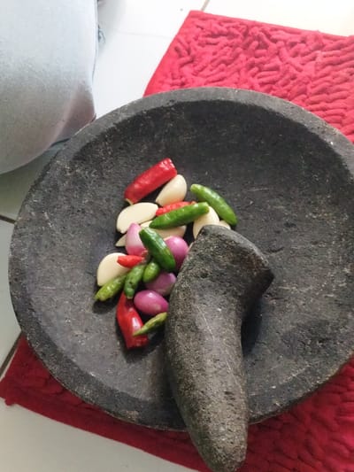 The image displays a close-up, overhead view of various fresh ingredients arranged within a dark grey stone mortar, commonly known as a "cobek" in Indonesia. A matching dark grey stone pestle, or "ulekan," rests vertically inside the mortar, positioned on the right side of the frame.
The ingredients gathered for preparation include a mix of bright red and green chilies, several peeled white garlic cloves, and small, purple-skinned shallots. These ingredients are typically used to create a spice paste or sambal, a staple condiment in Indonesian cuisine.
The mortar and pestle are placed on a red, textured mat or cloth. In the upper left corner, a light-colored fabric, possibly part of clothing, is partially visible, while a white surface (likely a floor or table) can be seen in the background, suggesting an indoor setting. The scene captures the initial stage of preparing a dish that requires grinding spices, a common activity in kitchens in Leuwiliang, Indonesia, consistent with the provided location context. No text is visible in the image, and the time of day or specific weather conditions cannot be determined.