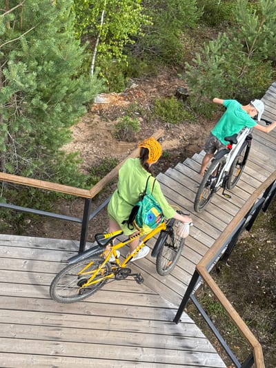 Two people with bicycles are on a wooden walkway in Peski, Russia. The person in the foreground is wearing a lime green outfit and a yellow headband, pushing a yellow bicycle. The person in the background is wearing a green t-shirt and patterned shorts, holding a white bicycle. The walkway is elevated and surrounded by trees and vegetation. The scene appears to be a park or recreational area.