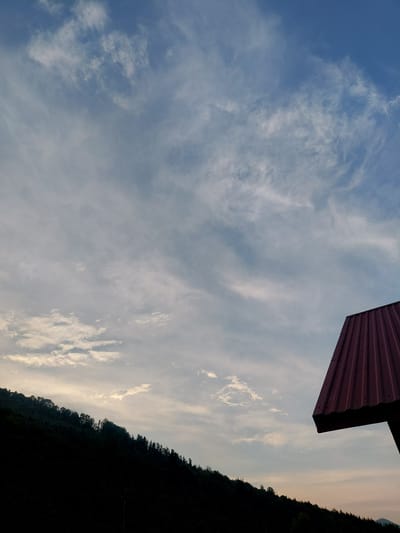This is a vertically oriented, wide shot of a cloudy sky and a dark, tree-covered hillside at what appears to be either sunrise or sunset. In the upper right corner of the image, a portion of a dark red, corrugated metal roof juts into the frame at an angle, suggesting the presence of a structure. The sky is a pale blue, with scattered wispy clouds that are illuminated from below by the setting or rising sun, creating a soft, ethereal glow. The clouds are more dense and brighter towards the right side of the frame, where they meet the horizon. The hillside on the right is almost entirely silhouetted against the sky, with only the faint outline of trees visible. This creates a stark contrast between the light and dark elements of the image. There is no visible text. The overall mood of the image is tranquil and serene, with a sense of natural beauty and isolation. The scene is likely set in a rural or natural environment, given the prominent hillside and the absence of any man-made structures besides the partial roof.