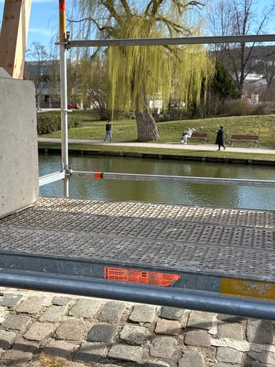 The image depicts an outdoor scene in Weißenburg in Bayern, Germany, under clear, sunny conditions. The foreground is occupied by a construction element featuring a concrete block on the left and a metal scaffolding platform extending across the lower half of the frame. The platform is gray, perforated, and features a red warning sticker with German text ("Original hat zum Layher Blitz-Gerüst ... Layher Absturzsicherung") on its horizontal support bar. Below the scaffolding, the ground is composed of irregular cobblestones.
Beyond the scaffolding and cobblestones, a calm body of water, green in tint, stretches across the midground. On the far bank of this water body, a grassy slope leads to a paved pathway. A large weeping willow tree with light green, early spring foliage stands prominently to the right of the scene's center. Three individuals are present on the pathway: one person in a dark jacket walks from left to right, one person in a white hoodie and dark pants sits on a wooden bench, and one person in a long dark coat walks from right to left, past two unoccupied wooden benches. In the background, beyond the pathway, are additional trees, some bare and some evergreen, and indistinct buildings under a clear blue sky.