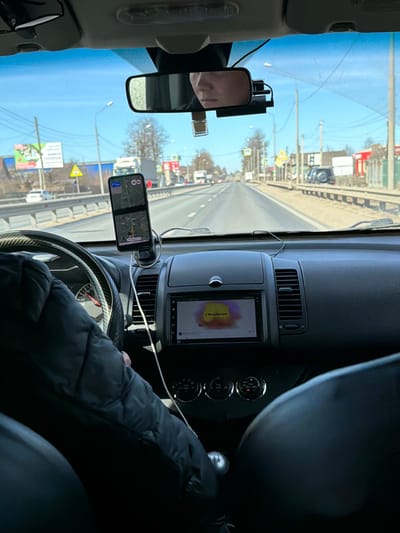 The image depicts the interior view from the passenger seat of a vehicle traveling northbound on a multi-lane highway in Zavidovo, Russia, under clear daylight conditions.
**Vehicle Interior:** The driver, whose face is partially reflected in the rearview mirror, has their left arm, clad in a dark quilted jacket, positioned on the steering wheel, which features a carbon fiber-textured cover. A smartphone, displaying a map navigation application with a green highlighted route, is mounted on the dashboard and connected via a white charging cable. Below it, the vehicle's infotainment system shows a colorful graphic with text partially legible as "Move this soul." A manual gear shift lever is visible in the lower center console. A dashcam is mounted near the rearview mirror.
**Exterior Environment:** The vehicle is on a paved road with at least two lanes in each direction, clearly marked by solid and broken white lines. A metal guardrail runs along the left side of the road. On the right shoulder, the ground is dry, with sparse brown vegetation and dirt. Utility poles with power lines line both sides of the road.
**Roadside Features:** On the left, a large billboard with Cyrillic text is visible, along with a white semi-truck in the distant opposing lane. On the right, a yellow triangular warning sign, green rectangular road signs, and additional utility poles are present. Further along the right side of the road, several low-rise commercial buildings with red roofs and white facades are visible. A dark-colored off-road vehicle is parked on the unpaved shoulder. The sky is clear blue, and trees in the background appear bare, suggesting late winter or early spring.
