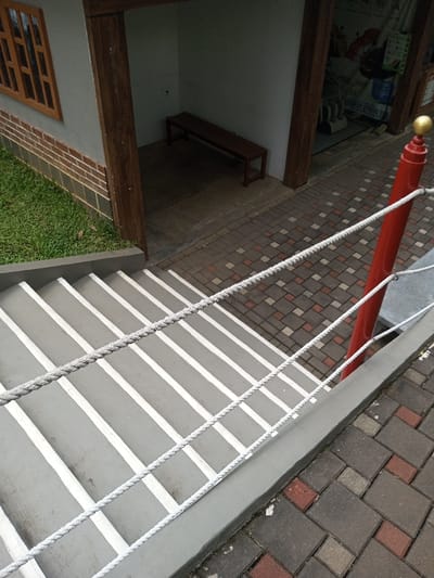 The image depicts an outdoor architectural scene in Tân Kỳ Commune, Vietnam, captured from an elevated perspective looking down. The primary subject is a descending gray concrete staircase with white painted lines on each tread. A light gray rope railing system, composed of three parallel strands, runs diagonally alongside the stairs, attaching to a prominent red cylindrical post topped with a gold spherical finial on the right. This post stands on a paved surface.
To the left of the staircase, a section of a building is visible, featuring a light gray wall above a brown brick base course. A brown wooden window frame with horizontal slats is set into this wall. Below an overhang of the building, a recessed alcove contains a dark brown wooden slatted bench against a light gray back wall. The floor of this alcove is concrete.
The ground around the building includes green grass to the left of the brickwork and a patterned walkway of interlocking concrete pavers in shades of gray and reddish-brown, situated at the top of the stairs and extending to the right. Beyond the recessed alcove, a dimly lit area is visible through an opening, revealing indistinct objects or equipment. No people or specific actions are depicted in the scene.