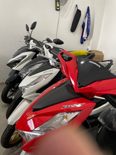 This image depicts a row of Honda Beat scooters parked indoors, likely in a showroom or storage area, in San Jose del Monte, Philippines. The scooters are various colors, including red, white, and black. They are arranged closely together, with their front ends facing the camera. The lighting is bright and even, suggesting an indoor environment. In the background, a white wall is visible, adorned with a black bag and a blue and white patterned shirt hanging on a hook. A box is also present in the background, possibly containing parts or accessories. The scooters appear to be new and in good condition. The overall scene conveys a sense of organized storage or display of vehicles.