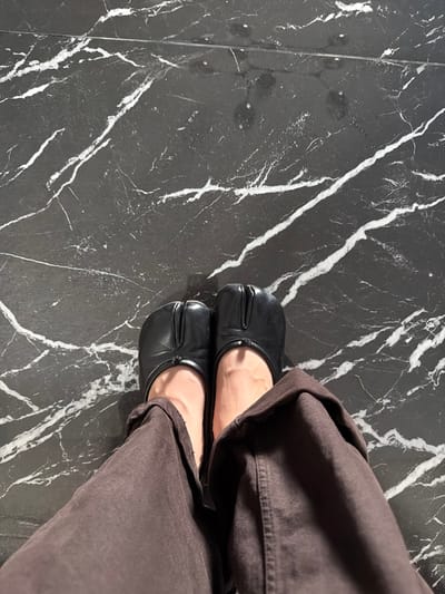 The image presents a top-down, close-up view of a person's lower legs and feet, located indoors on a polished black marble floor in Villahermosa, Mexico. The individual is wearing black leather tabi-style shoes, characterized by a split-toe design. Dark brown trousers with a relaxed fit cover the calves, revealing bare ankles and the top of the feet. The floor consists of large, reflective black marble tiles prominently patterned with irregular white veining. Overhead lighting illuminates the scene, casting subtle reflections on the shoes and floor surface. No other subjects or actions are discernible.