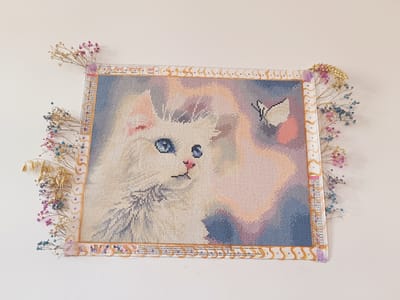 The image shows a diamond painting of a white cat with blue eyes, a butterfly, and a pinkish-orange background. The painting is framed with a decorative border and decorated with dried flowers. A color list and instructions in German are visible on the right side of the painting. The painting is hung on a white wall. This image was taken in the city of Legnica, Poland.