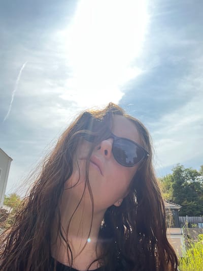A low-angle, upward-facing photograph captures a subject, likely female, with long, wavy, dark brown hair that appears damp, obscuring parts of her face. She wears dark-framed sunglasses with reflective lenses, partially revealing her nose and lips. A small, reflective stud earring is visible in her right earlobe. The upper portion of the image is dominated by a bright, partly cloudy sky with a prominent sun glare near the top center, and a faint contrail is visible in the upper left. In the background, on the left, a portion of a light-colored modern building is visible. On the right, beyond green foliage, a building with a reddish-brown tiled roof and light-colored walls is partially seen, alongside tall green grass or reeds. The scene is situated in Bad Sobernheim, Germany.