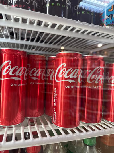 The image captures a close-up, angled view of beverages displayed horizontally in a refrigerated unit, likely in a commercial setting such as a store in Donets, Ukraine. The main subjects are multiple rows of red Coca-Cola cans, stacked on white plastic shelving. These cans prominently feature the white "Coca-Cola" logo and text like "ORIGINAL TASTE" and "ORIGINAL FINAL TASTE".
To the left, partially obscured by the shelving, are black Monster Energy drink cans, showing parts of their "M" logo and "ENERGY" branding. The white plastic shelves appear cold, possibly with some frost or condensation, indicating the drinks are chilled. Faint reflections of indoor lighting are visible on the glossy surface of the cans. In the background and to the right, other plastic bottles with green and red caps can be vaguely seen.
Notable visible text includes "Coca-Cola", "ORIGINAL TASTE", "ORIGINAL FINAL TASTE", and on the sides of some Coca-Cola cans, "СОРТУЙ УПАКОВКУ" (Ukrainian for "sort packaging") and "SORTEAZA AMBALAJUL" (Romanian for "sort packaging"). Text like "MONSTER ENERGY" and "ENERGY" is also visible on the Monster cans. No individuals are present in the frame.