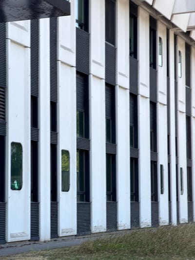This is a vertical, eye-level, outdoor, close-up shot of the exterior of a modern building, likely a multi-story office or residential complex, in Rungis, France. The building features a repeating pattern of white horizontal panels interspersed with dark, ribbed sections that appear to be vents or grates. Within these dark sections, there are horizontal, elongated windows that offer glimpses of the interior, reflecting the surrounding environment. The architecture is sleek and contemporary, with clean lines and a minimalistic aesthetic.
The image captures the building from a low angle, looking upwards. The sunlight suggests it is daytime, and the lighting indicates it is likely overcast or the sun is not directly hitting the facade. The overall impression is one of ordered repetition and functionality. The texture of the ribbed sections contrasts with the smooth, white panels, creating visual interest.
To the right of the building, a strip of green grass is visible, suggesting the building is situated in an area with some landscaping, possibly a park or a corporate campus. The image is slightly blurred, particularly in the foreground and background, with the building's facade in sharper focus. No people are visible in the image, and the focus is solely on the architectural elements of the building. There is no visible text in the image.