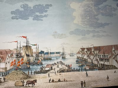 This detailed image depicts a bustling harbor scene under a partly cloudy sky. The light blue sky is populated with scattered grey and white cumulus clouds. The water in the harbor is a calm, blue-green.

In the foreground, a wide, light-colored quay is busy with numerous figures. On the left, a large, irregularly stacked pile of light-colored timber or wood dominates the ground. Adjacent to this, a brown horse pulls a cart laden with a large, textured, round bundle or sack. Several individuals are present in this area, including one in red on a small boat near the wood pile. In the mid-foreground, three figures are seated on the ground, seemingly engaging in commerce with several baskets placed before them. Various other figures, including children, are scattered across the quay, walking or standing.

To the left, multi-story white wooden buildings with reddish-brown gabled roofs line the waterfront. Docked alongside these buildings are several large, three-masted sailing ships with sails partially furled or lowered. Each of these ships prominently displays Danish flags (red with a white Scandinavian cross) on their masts.

In the center of the image, a long, narrow wooden pier extends into the harbor, densely populated with small figures engaged in various activities. A small, simple building with a red roof stands at the base of this pier. To the right of this main pier, a shorter pier or dock is also visible, with boats tied alongside and more figures.

The right side of the harbor features more multi-story white wooden buildings with steep, reddish-brown roofs, some built directly over the water on stilts or pilings. Several more sailing ships and smaller vessels are moored here, some with their sails furled, others appearing to be in various stages of loading or unloading.

In the background, more ships and distant buildings with blue roofs can be seen, indicating the extent of the port. The overall scene conveys a sense of active trade and daily life in a northern European port, characterized by its distinctive architecture and the ubiquitous presence of Danish maritime flags.