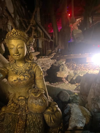 A gold statue is located in Baan Don Sai, Thailand. The statue is situated in a cave-like environment with rocks and root-like structures surrounding it. The area above appears to be a constructed ceiling or overhead structure with red and white lights.
