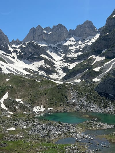 This image captures a high-alpine landscape in Shalë, Albania, under a clear, cloudless blue sky. Prominent in the background are jagged, grey mountain peaks extensively covered with snow patches, particularly on their upper and mid-slopes. Large, irregular fields of snow are visible on the steep, rocky terrain, interspersing with exposed rock formations and areas of green vegetation. In the mid-ground, a distinct turquoise-green alpine lake with several small, rocky islands and clear water reflects the sky. Below this main lake, smaller, shallower pools of water and marshy areas are interspersed with abundant grey rocks and sparse green growth. On a snow-covered slope on the left side of the image, multiple small, dark figures, consistent with hikers, are discernible traversing the terrain. The environment is rugged, characterized by rock, snow, and nascent vegetation, indicative of a post-winter or early summer period.