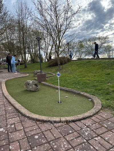 This is an outdoor scene at a miniature golf course. The focus is on hole number 15, indicated by the number on a pole near the hole. The hole is circular, with a green surface and a stone border. There is a rock obstacle on the putting surface. Adjacent to the hole is a paved area with brown bricks. In the background, there are people, trees, and a grassy area. The sky is overcast. The location is Gunzenhausen, Germany. People are engaged in playing miniature golf.