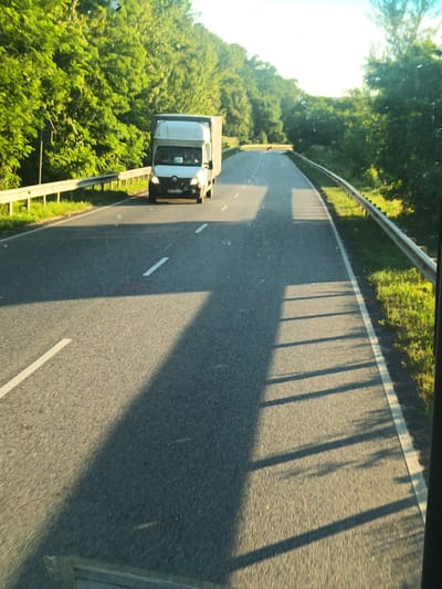 A white panel van with a box trailer is traveling on a two-lane asphalt road lined with guardrails and trees. The van is in the left lane, with its headlights on. The sun is shining brightly, casting long shadows across the road. The scene appears to be a rural area, possibly on the outskirts of a city. The specific location in Kamianytsia, Ukraine cannot be determined from the image alone.