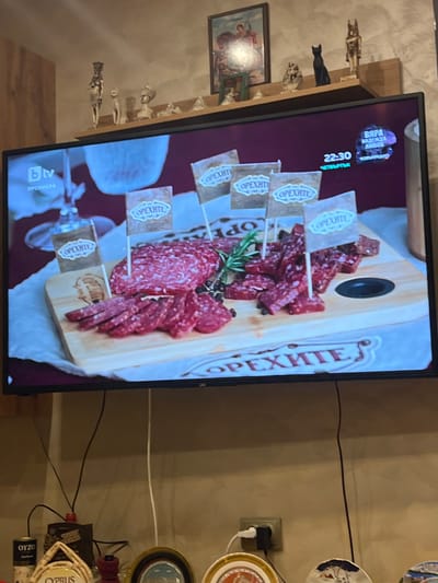 An indoor scene, possibly in Popintsi, Bulgaria, features a flat-screen television displaying a food advertisement or cooking segment, viewed at an angle. The TV screen shows a wooden serving board laden with slices of cured meat, likely salami or sausage, garnished with fresh rosemary and whole black peppercorns. Small decorative flags, bearing the brand name "ОРЕХИТЕ" (OREHITE) in Cyrillic, are stuck into the meat portions. The same brand name appears in a larger, stylized font along the right edge of the wooden board. In the top left corner of the screen, the "bTV" channel logo is visible, alongside "22:30" indicating the time, and "ЧЕТВЪРТЪК" (THURSDAY). Additional smaller text is present in the top left, which is partially obscured.
To the left of the television, a wooden shelf holds several decorative figurines, including Egyptian-themed statues such as Anubis, a black cat, and other smaller, indistinct figures. Below the shelf, a framed picture hangs on the wall. To the right of the TV, the wall features an electrical outlet with a power adapter plugged in. Above and below the outlet, multiple decorative plates hang on the wall. Further down, various cables are visible, along with small bottles or cans, one of which clearly shows "OYZO" and another "CYPRUS." The overall setting suggests a domestic environment, possibly a living or dining room, with various personal decorations. The time indicated on the screen, 22:30, points to late evening.
