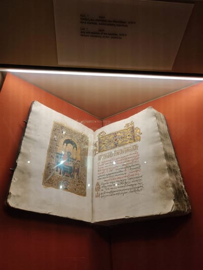 The image displays an open, ancient illuminated manuscript inside a glass display case. The manuscript is open to a page featuring a detailed miniature painting on the left-hand side, depicting a figure seated at a desk. The right-hand page contains densely written text in red and black ink, adorned with ornate gold lettering and decorative borders. The manuscript rests on a light-colored surface, with the surrounding display case having an orange-red textured backdrop. Above the display case, a label provides information in both Greek and English, indicating the manuscript is from the year 1618/9 and is from the Varlaam monastery. The Varlaam monastery is located in Kalambaka, Greece, known for its monasteries perched atop rock formations. The lighting within the display case creates reflections on the glass, slightly obscuring the details of the manuscript.