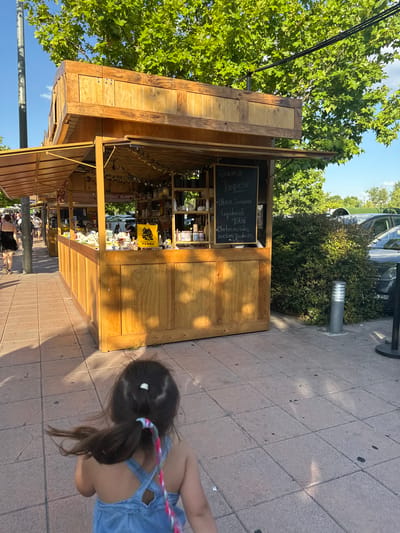 A wooden kiosk, likely a market stall, is positioned outdoors on a paved walkway. The kiosk is filled with various products displayed on shelves, and a chalkboard menu is visible on its facade. To the right of the kiosk, dense green bushes and parked cars are present. In the foreground, a young child with dark hair in a ponytail, wearing a blue denim dress, is walking away from the camera. The child's hair is being blown by the wind. The scene is illuminated by sunlight, casting shadows on the ground. The environment suggests a public outdoor space, possibly a park or a shopping area, given the presence of cars and other people walking in the background. The text on the chalkboard menu, visible in the crop, includes "GRACIAS!!! PARQUESUR" and promotional messages like "ULTIMA SEMANA LIQUIDACIÓN TOTAL OFERTAS en todos nuestros productos." This suggests the location is likely within the Parquesur complex in Leganés, Spain, as indicated by the text on the chalkboard and the overall setting.