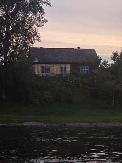 A weathered, wooden house with a dark, corrugated roof stands on a grassy embankment overlooking a body of water. The house has yellow siding with circular insulation elements visible and multiple windows, one with a blue frame. Dense green foliage surrounds the structure, partially obscuring it. The water in the foreground is dark and reflects the soft light of a pale, cloudy sky. A large tree with dark leaves is silhouetted against the sky on the left side of the frame. The scene appears to be a rural setting in the village of Yukseyevo, Russia, during twilight.