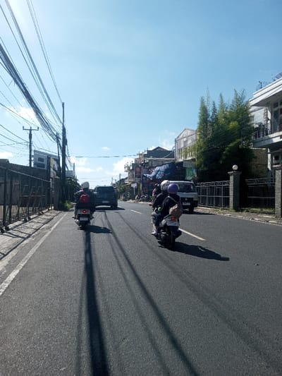The image depicts a daytime street scene in Ban Phonhxavanh, Laos, characterized by clear weather and bright sunlight. The perspective is from slightly above ground level, looking down a paved, two-lane asphalt road. The road surface exhibits a solid white line marking its left edge and a partial yellow line visible on the right. Long, dark shadows, likely cast by overhead utility wires and poles, stretch across the asphalt from the left side of the frame towards the right.

Two motor scooters are prominent in the foreground and mid-ground, traveling away from the viewer. The scooter on the left carries a single rider wearing a white helmet and a dark top, possibly with a red backpack. The scooter on the right carries two individuals; the front rider wears a dark top and a dark-colored helmet, while the passenger behind them also wears a dark top and a light-colored helmet, carrying a light-colored bag on their back. Both scooters are dark in color. A dark-colored SUV or similar vehicle is visible further down the road, behind the left scooter. Ahead of the right scooter, a light-colored van or small truck is parked or moving slowly on the right side of the road.

On the left side of the road, a dark-colored metal fence runs parallel to the roadway, behind which a utility pole supports a dense network of black electrical and communication wires. Buildings are partially visible behind the pole and fence. On the right side of the road, a low wall constructed from stone and capped pillars with black metal fencing is present. Behind this fence, several tall, green bamboo trees stand next to a multi-story building featuring balconies. Further down the street on the right, a row of commercial buildings with various signs, awnings, and blue tarpaulins are visible, indicating retail or service establishments. The sky above is clear and bright blue with scattered faint white clouds.