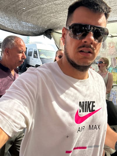 The media file shows a group of people in an outdoor market setting, possibly El Parador de las Hortichuelas, Spain. The foreground is dominated by a man wearing sunglasses and a white t-shirt with a large "NIKE" logo and "AIR MAX" text printed on it. He is looking towards the camera. Behind him, another man with graying hair is visible, looking upwards. Further in the background, a woman with blonde hair and sunglasses is looking to her left with an open mouth, suggesting she is speaking or surprised. There are clothing items displayed in the background, suggesting a market stall. The lighting suggests it is daytime, with bright sunlight casting shadows. The overall scene appears casual and candid.
