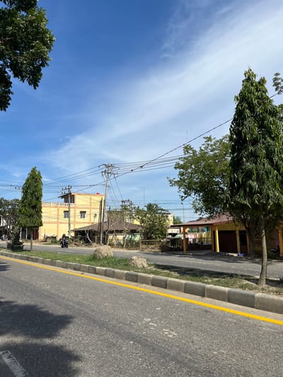 A street scene in Tanahterban, Indonesia shows a paved road with a bright yellow line marking the edge of the lane. A concrete curb with grass and several large rocks runs alongside the road. Buildings with yellow walls and orange trim line the street, interspersed with trees and utility poles with numerous wires. A single motorcycle is visible on the road, with a rider wearing a helmet. The sky is blue with scattered white clouds.