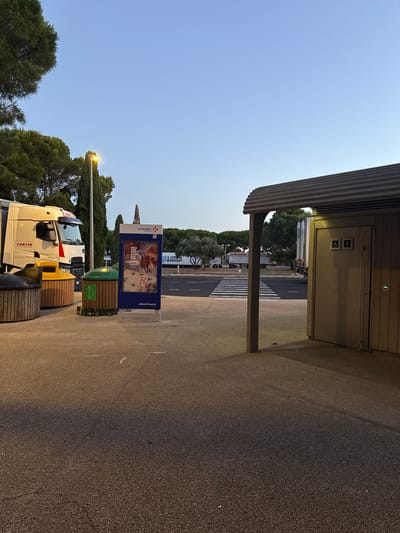 This image depicts a rest stop area, likely along a highway or major road, in or near Florensac, France. In the foreground, a paved area is visible with several waste receptacles, including two wooden-clad bins and one yellow bin. A green bin with a bottle recycling symbol is also present, with discarded glass bottles around its base. To the left, the front of a white semi-trailer truck with "CURCIO" visible on its side is parked.

A digital advertisement board with the "VINCI Autoroutes" logo stands next to the bins. The advertisement features imagery of playing cards and the text "#BIENVOYAGER." Behind the advertisement, a crosswalk is marked on a paved road, with several semi-trailer trucks parked and moving in the background.

On the right side of the image, a modern restroom facility with a corrugated metal roof is visible. The entrance to the facility has two signs above it: one depicting a wheelchair user, and another a standard male/female symbol. The exterior walls of the restroom are made of vertical wooden planks. The environment appears to be an open area with trees and clear sky, suggesting it is daytime.