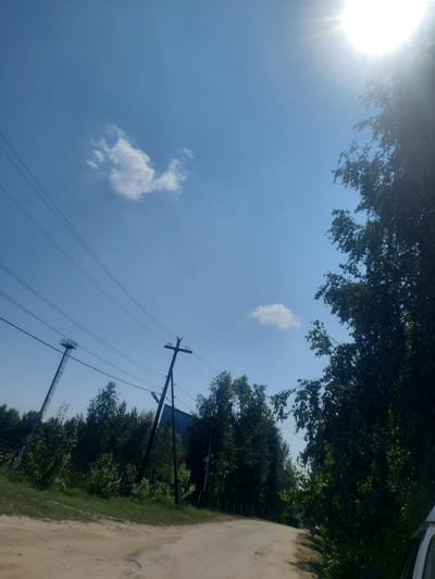 The image depicts a rural scene under a clear blue sky with a bright sun in the upper right corner, casting lens flares and highlighting the foliage of a dense tree on the right. In the foreground, a dusty dirt road curves gently into the distance. Power lines stretch across the sky, supported by utility poles. One pole is a tall, lattice-like structure with a platform at the top, located on the left side of the image. Another, simpler pole with crossbars is visible further to the right. The road is bordered by lush green vegetation, including grass and trees, on both sides. In the background, a large, dark, angular building can be seen through the trees. The overall impression is one of a bright, sunny day in a somewhat remote, possibly industrial or semi-rural area. The specific location of Pavlovsk, Russia, is not visually identifiable from the image itself.