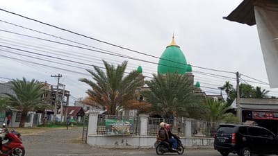 In the city of Geulanggang Teungoh, Indonesia, a street scene unfolds under an overcast sky. A prominent feature is a large, green domed building, likely a mosque, with smaller green domes and a golden spire. Palm trees, some with dry fronds, are scattered in front of the building, and a metal fence encloses the property. A recruitment banner for Tentara Nasional Indonesia is visible on the fence. On the street, a black SUV is parked on the right, and a red motorcycle is partially visible on the left. A two-person motorcycle passes by, with a rider and passenger wearing helmets. Other individuals are seen walking on the sidewalk and in the background, near buildings and what appears to be a utility pole with an electrical transformer. The road is paved, and a gravel area is visible on the left side of the street.