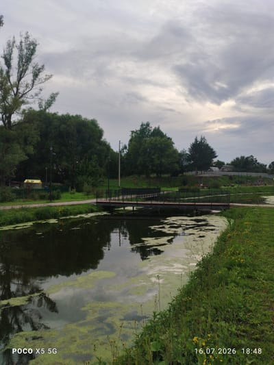 A landscape scene depicts a canal or river with a small footbridge crossing it. The water is dark and partially covered with green algae. On the left bank, there is lush green vegetation, including trees, and a small yellow building with a fence. On the right bank, a grassy embankment rises, with more trees and some distant buildings visible. A pathway runs along the left bank. The sky is overcast with heavy clouds. The image is timestamped "16.07.2026 18:48" and branded "POCO X5 5G". The location is identified as Aksubayevo, Russia. There are no people visible in the immediate foreground or on the bridge.
