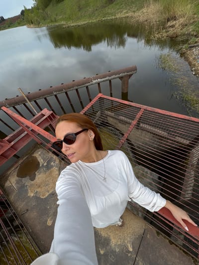 A woman wearing sunglasses and a white long-sleeved shirt stands on a rusty metal structure with a red grid, overlooking a body of water. The water reflects the cloudy sky. Green vegetation is visible on the banks. The location is likely Nozhovka, Russia.