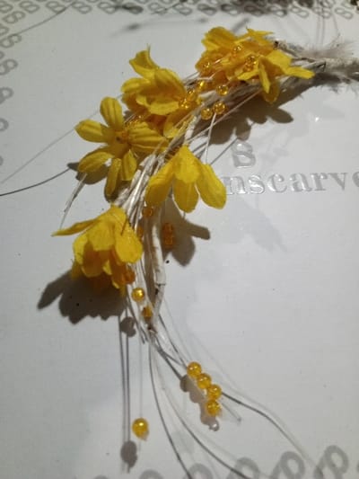 A decorative floral arrangement sits on a white surface. Yellow fabric flowers are clustered on thin, white stems interspersed with small yellow beads. The stems extend downward, some ending with a single bead. A feathery accent sits near the top of the arrangement. Shadows are cast on the surface, which has embossed lettering reading "nscarved".
