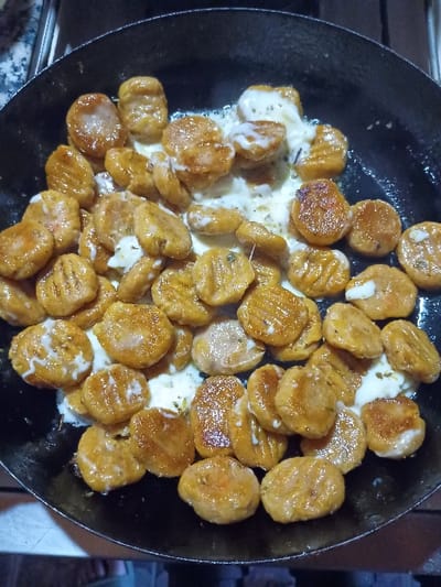 The image shows a black frying pan filled with cooked gnocchi. The gnocchi is coated in a white sauce, likely a creamy cheese sauce. The pan is sitting on a stovetop in a residential kitchen in El Talar, Argentina. The coordinates provided, -34.473 latitude and -58.639 longitude, correspond to a residential area in El Talar. The gnocchi appears to be cooked and ready to be served. The scene suggests a simple meal preparation in a home setting.