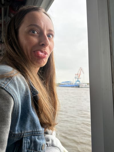 This close-up selfie captures a young woman with a playful expression, her head tilted to the right within the frame. Her long, light brown hair cascades around her shoulders. She looks directly into the camera with wide blue eyes, her tongue slightly extended in a humorous gesture. She is dressed in a blue denim jacket layered over a grey hooded garment.

Behind her, an outdoor scene is visible through what appears to be a window. The sky above is bright and overcast, suggesting daytime on a cloudy day. A large body of water, likely a river or bay, stretches across the middle ground. In the distance, an orange industrial crane and other structures hint at a port or waterfront industrial area, consistent with the context of Nizhneleninskoe, Russia. A light-colored window frame or sill is visible along the top edge of the image. The overall mood conveyed is spontaneous and lighthearted. There is no visible text.