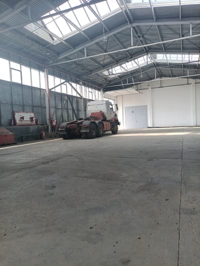An interior view of a large, industrial warehouse in Aksay, Kazakhstan, reveals a sparsely populated space dominated by concrete flooring and a corrugated metal roof with skylights. A semi-trailer truck, likely used for hauling, is parked in the center of the frame, facing away from the camera. The truck's chassis is visible, with its tires and rear wheels prominent. To the left of the truck, a red metal object, possibly a container or bin, rests on the ground. Further left, against a gray metal wall, is a white sign with red lettering that reads "ПОЖАРНЫЙ ЩИТ №1" (Fire Shield No. 1). Two fire extinguishers are also visible on the wall. The warehouse features tall windows along one wall, allowing natural light to enter, and a large, closed bay door at the far end of the space. The overall impression is one of a functional, empty industrial setting.