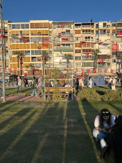 The image depicts a public park in İzmir, Türkiye, located at the coordinates 38.438 latitude and 27.141 longitude. The scene features a grassy area, a large, multi-story building, and a metal trellis structure. People are walking and interacting in the park. Some are seated on a bench within the trellis, while others are sitting on the grass. Shadows cast from the setting sun stretch across the grass. Several Turkish flags are draped across the building's facade.