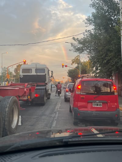 The image depicts a street scene in Gerli, Argentina, viewed from a stopped vehicle at what appears to be a traffic light. The sky is partly cloudy, with a visible rainbow arc in the upper right quadrant. Multiple traffic lights are illuminated red, indicating stopped vehicular movement.

In the immediate foreground, a portion of a vehicle's dashboard and windshield wipers are visible, with reflections of red taillights from the vehicle ahead.

Directly in front, in the right lane, is a red Hyundai Atos GLS hatchback equipped with a roof rack. Its Argentinian license plate reads "DCA 072," and its brake lights are illuminated. Reflections of foliage are visible on its rear window. Behind this vehicle, a silver sedan is partially visible.

In the adjacent left lane, a white and dark grey semi-truck cab with an empty red flatbed trailer is stopped. A circular decal displaying "80" is present on the rear of the truck cab. Its brake lights are also illuminated. Ahead of the semi-truck, a person on a motorcycle, wearing a red jacket and carrying a red delivery box featuring a white "P" logo, is stopped.

The road surface is asphalt. Utility poles with overhead wires, streetlights, and green-leafed trees line both sides of the street. Buildings are partially visible in the background. The lighting suggests late afternoon conditions.