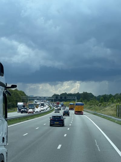 The image shows a congested multi-lane highway captured from the perspective of a vehicle, likely a truck, as indicated by the side mirror and part of the cabin visible in the bottom right corner. The highway stretches from the foreground into the distance, curving gently to the left.

The sky above is dramatic and overcast, with a clear division between dark, heavy grey clouds dominating the left side of the frame and lighter, brighter grey clouds on the right, suggesting dynamic weather conditions. Lush green trees and vegetation line the sides of the highway, forming a dense barrier between the road and the distant landscape.

Traffic is heavy, particularly in the left lanes, where numerous cars, vans, and trucks are bumper-to-bumper, appearing either stationary or moving very slowly. In the foreground and middle ground of the rightmost lane, there are a few vehicles, including two trucks carrying large, bright yellow covered loads, and several cars. One dark blue car in the right lane prominently displays the Dutch license plate "N-864-OJ," confirming the location context of Bathmen, Netherlands. Overhead gantries with various signals or signs span across the highway in the mid-distance. The scene depicts a typical daytime traffic jam under a moody sky.