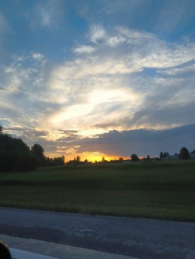 A sunset over a rural landscape in Barlak, Russia. The sky is filled with dramatic clouds, transitioning from deep blues and grays to bright oranges and yellows near the horizon. A bright sun is partially obscured by clouds, casting a warm glow over the scene. In the foreground, a dark green field stretches out, dotted with trees. The silhouette of buildings can be seen in the distance. A grey asphalt road runs along the bottom of the frame, suggesting a roadside view.