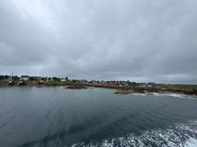 The image shows a view of the Sleneset area in Norway from the water. A small coastal village with several red and white houses is visible on the shore. The houses are situated on rocky terrain with some greenery. The sea in the foreground is dark and choppy, with white foam indicating motion, possibly from a boat. The sky is overcast with heavy gray clouds, suggesting a cloudy or possibly rainy day. The scene is natural and depicts a typical northern coastal landscape. There are no people visible in the image.