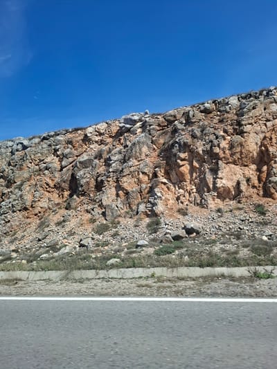 The image depicts a rocky cliff face adjacent to a road, with a clear blue sky visible above. The cliff is composed of various sized rocks and stones in shades of grey and brown. There are small patches of vegetation growing on the cliff face and along the roadside. The road is paved and marked with a white line. The location is Bogyovtsi, Bulgaria, based on the available information. The scene contains no people, subjects, objects, actions, interactions.